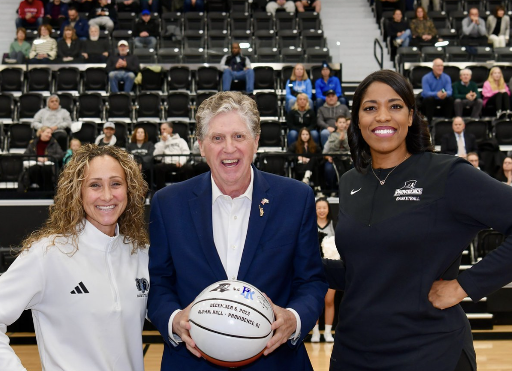 URI head coach Tammi Reiss, Rhode Island governor Dan McKee and Providence head coach Erin Batth pose for a photograph. McKee is holding a commemorative basketball in recognition of the state rivalry game.